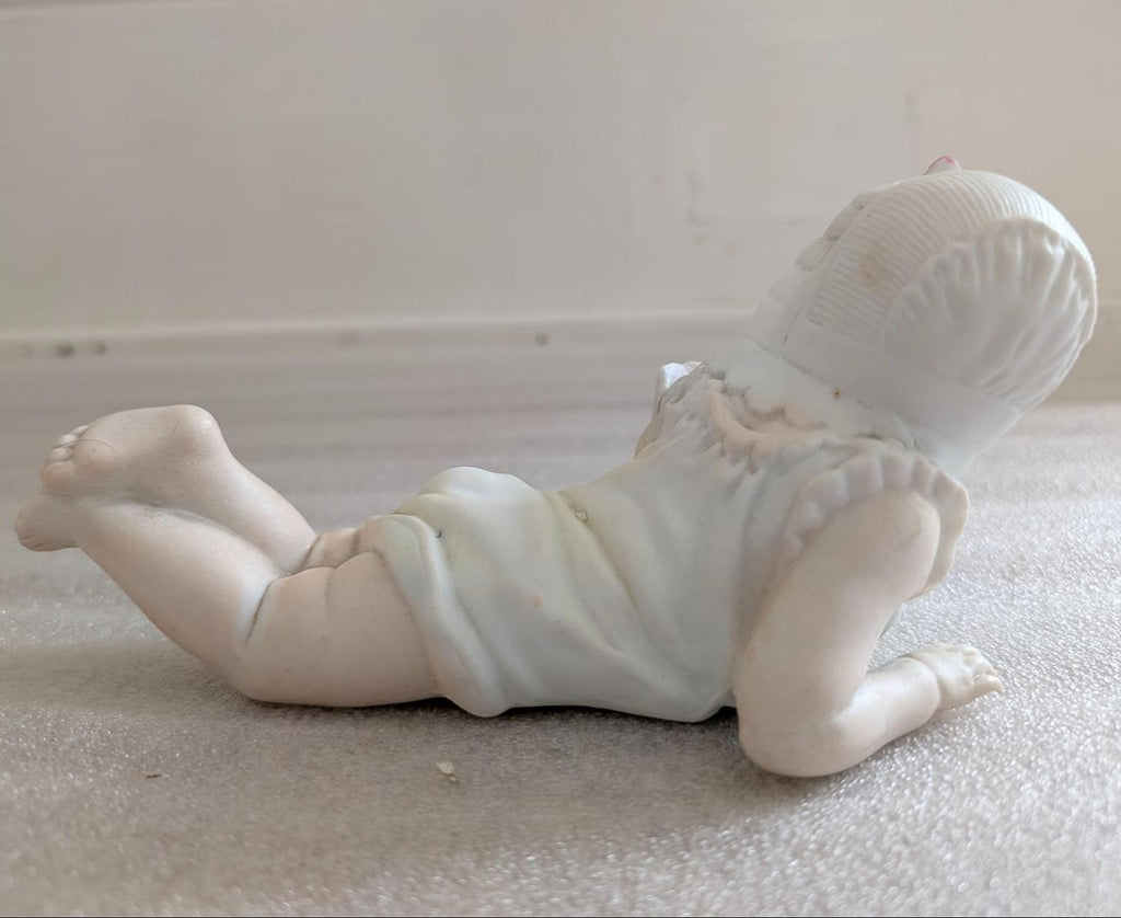Porcelain baby figurine reclining on belly, wearing bonnet and white romper