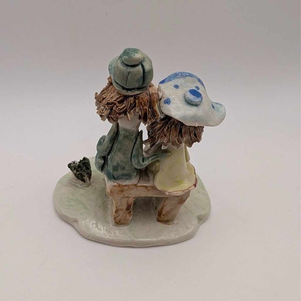 Small ceramic figurine of two friends, one in a cap and green dress, on a rounded base