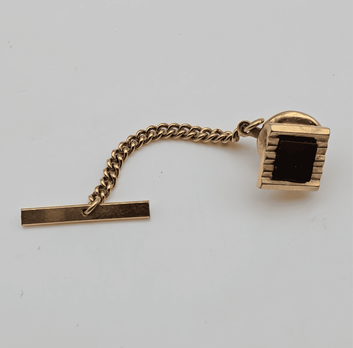 Gold-tone tie tack pin with geometric dark stone, connected chain and bar clasp