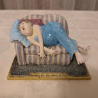 A ceramic figurine of a woman sleeping on a striped chaise lounge, displayed on a textured base with the phrase 'Waiting for Her Nails to Dry'.