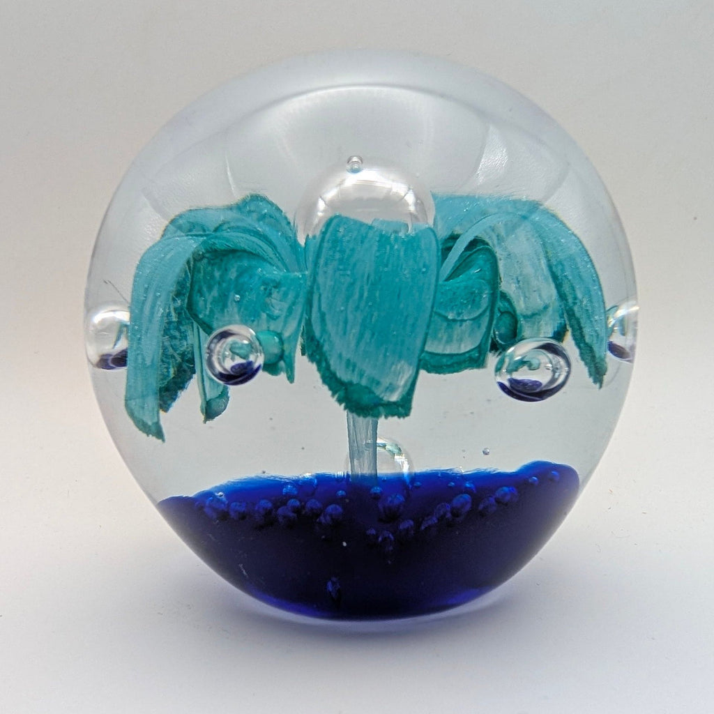 A clear glass paperweight shaped like a globe, featuring a painted teal palm tree on a dark blue base with small bubble details inside, set against a plain light gray background.