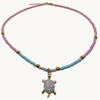 A beaded necklace with pink and light blue beads, accented with small gold spacer beads and a central gold turtle charm decorated with pink and blue mosaic details, shown on a plain light gray background.