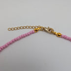 Close-up of a beaded necklace with light pink faceted beads, gold-toned chain extender, lobster clasp, and small gold beads on a plain light background.