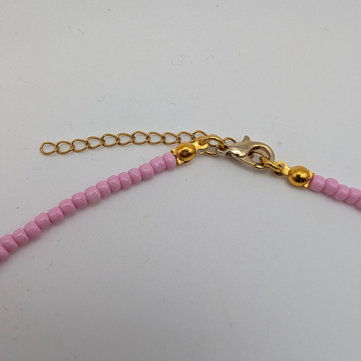 Close-up of a beaded necklace with light pink faceted beads, gold-toned chain extender, lobster clasp, and small gold beads on a plain light background.