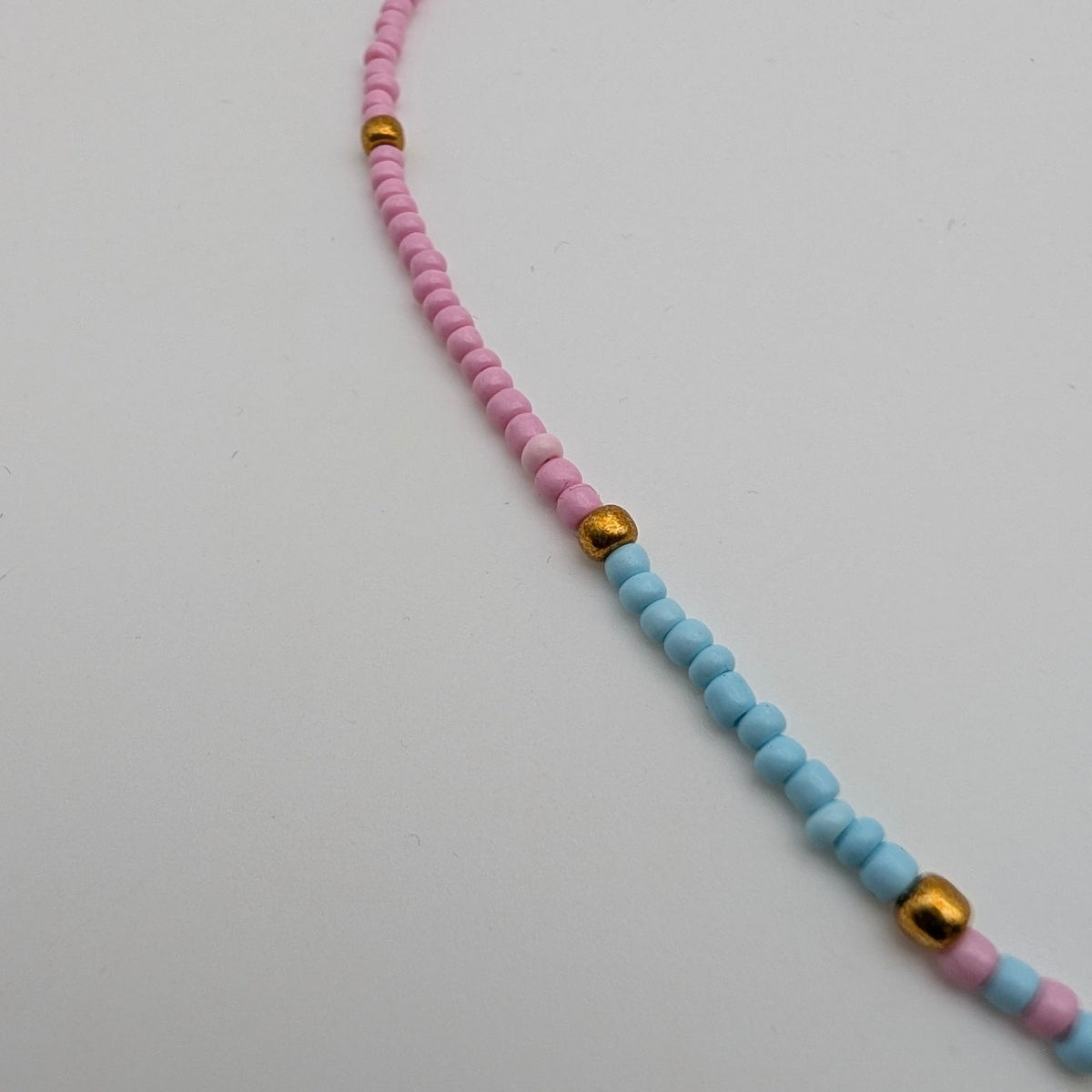 A beaded necklace with alternating strands of pink and light blue beads, accented by small gold spacer beads and a gold-tone clasp, shown on a plain light gray background.