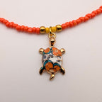 A beaded necklace with small orange-red beads, gold-toned spacer beads, and a central turtle charm decorated with orange, blue, white, and gold enamel.