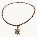 A beaded necklace with alternating red and green beads, featuring a gold-tone turtle pendant with a colorful floral inlay, displayed on a light gray background.