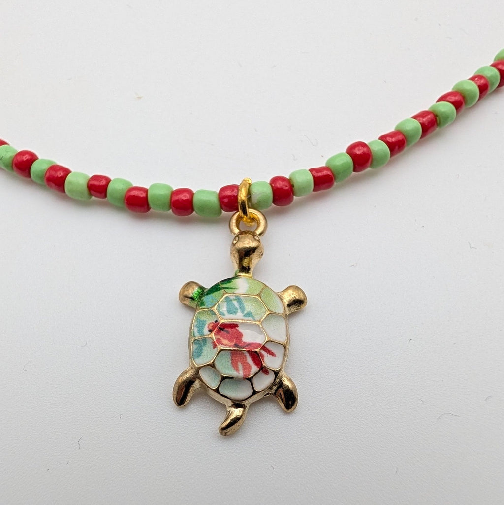 A beaded necklace with alternating red and light green beads and a gold turtle pendant featuring a colorful floral enamel design, displayed on a plain white background.