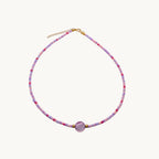 A delicate beaded necklace with small pink, purple, and white beads, featuring a central round light purple stone and a gold-tone clasp with an extender chain, displayed on a plain white surface.