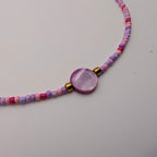 A beaded necklace featuring small pastel pink, purple, and white beads with a central round purple shell-like pendant and small gold spacer beads, displayed on a light gray surface.