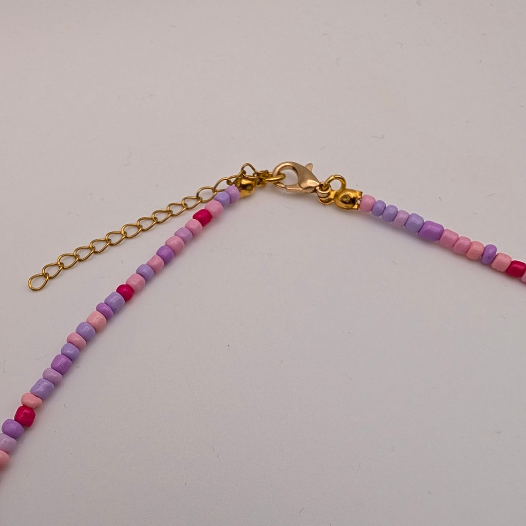 A close-up of a beaded chain accessory with small round beads in pink, purple, and peach tones, connected by a gold-tone chain extender and lobster clasp, on a plain light background.