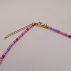 A close-up of a beaded chain accessory with small round beads in pink, purple, and peach tones, connected by a gold-tone chain extender and lobster clasp, on a plain light background.