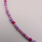 A strand of small faceted beads in pastel pink, lavender purple, and bright magenta pink, arranged diagonally on a plain off-white surface.