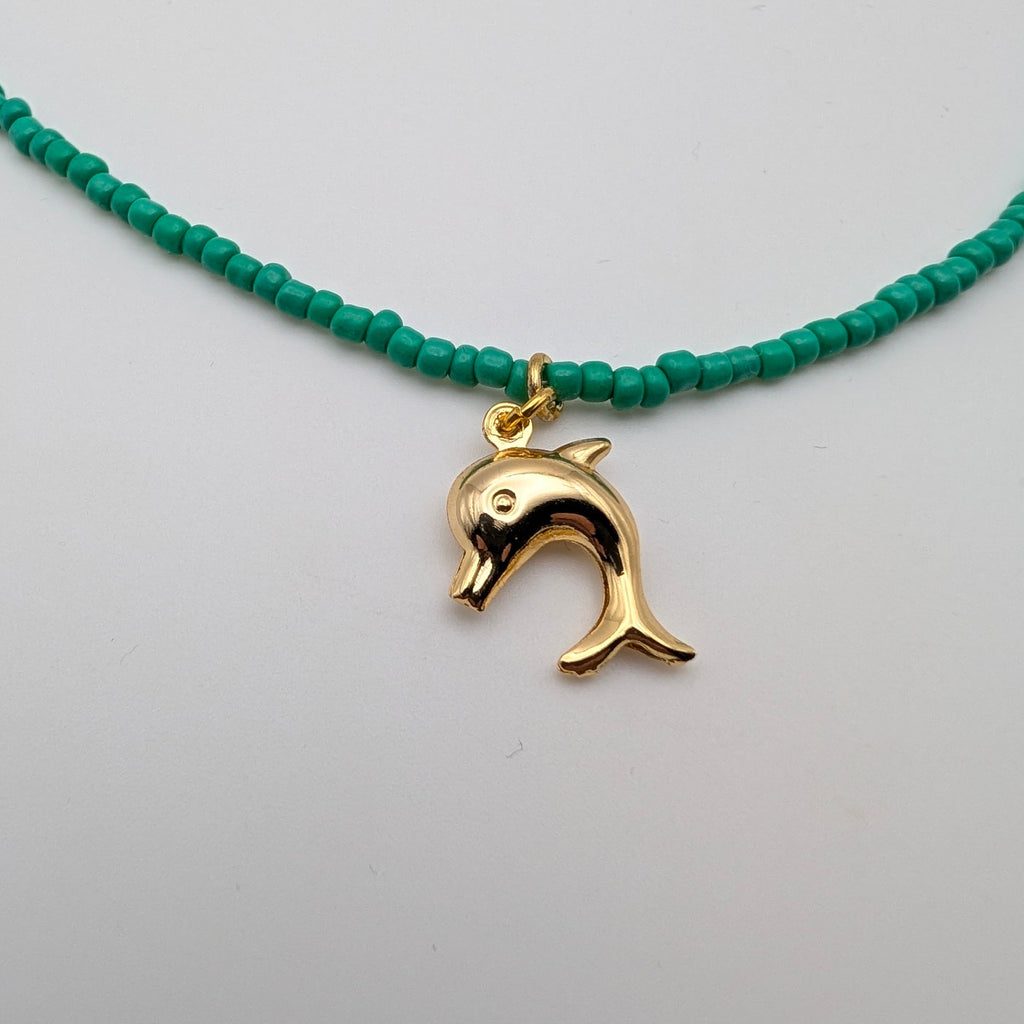 A beaded necklace featuring small, round turquoise-green beads and a polished gold-tone dolphin charm, displayed against a clean white background.