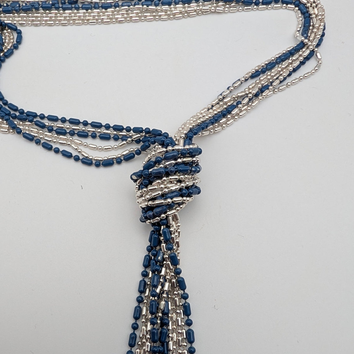 A multi-strand beaded tassel necklace featuring alternating rows of dark blue and silver seed beads, arranged on a plain light gray background.