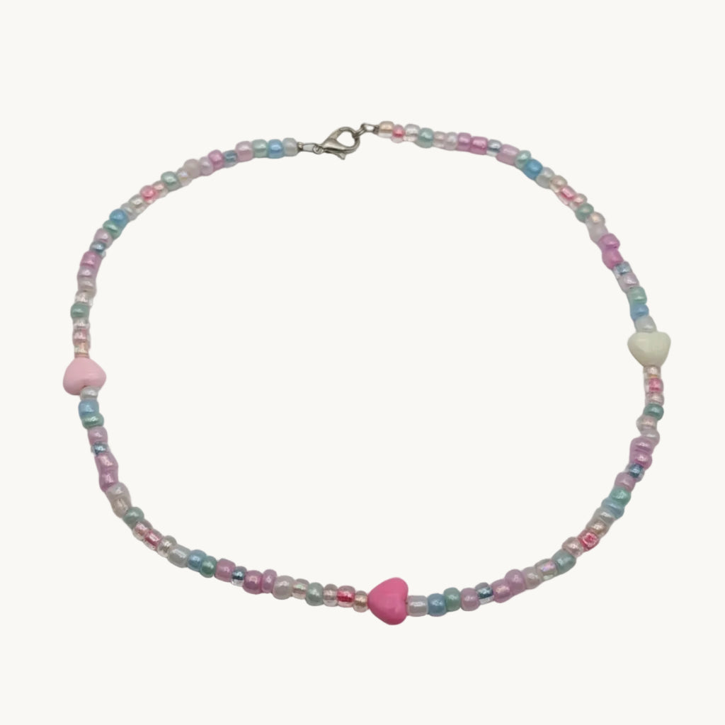 A delicate beaded necklace with pastel pink, light blue, and white beads, featuring a central pink heart-shaped bead and a silver clasp, displayed on a plain light gray background.
