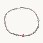 A delicate beaded necklace with pastel pink, light blue, and white beads, featuring a central pink heart-shaped bead and a silver clasp, displayed on a plain light gray background.