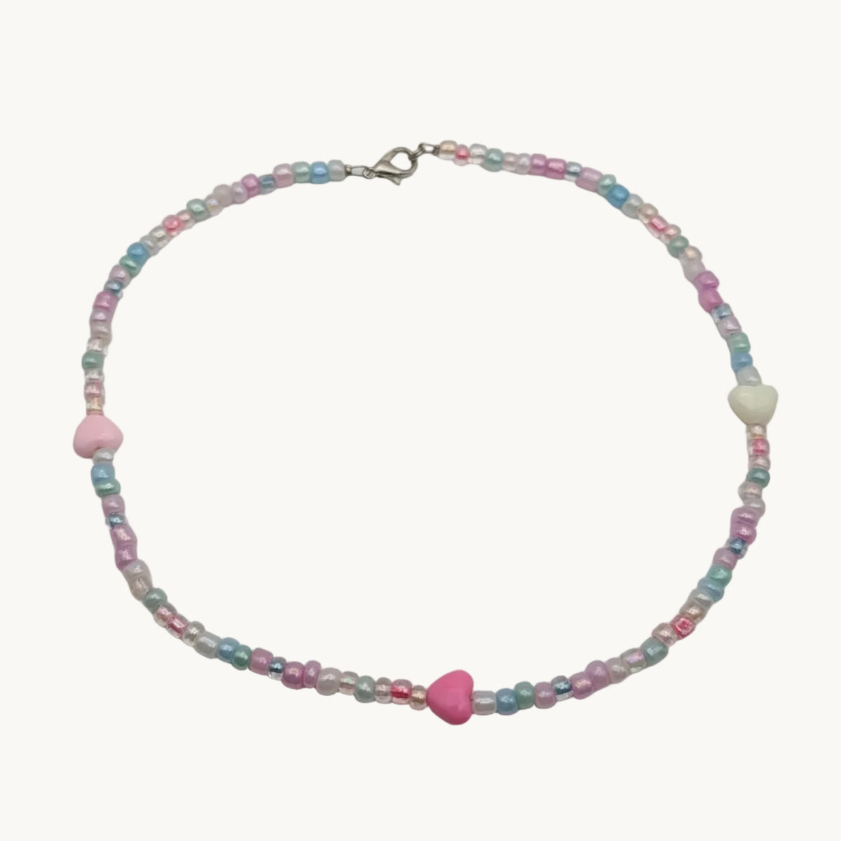 A delicate beaded necklace with pastel pink, light blue, and white beads, featuring a central pink heart-shaped bead and a silver clasp, displayed on a plain light gray background.