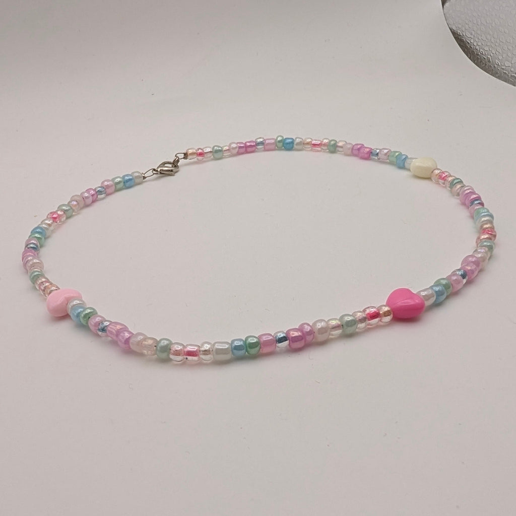 A pastel beaded necklace with pink and blue beads, a small pink heart charm, and a silver clasp, displayed on a light gray surface.