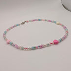 A pastel beaded necklace with pink and blue beads, a small pink heart charm, and a silver clasp, displayed on a light gray surface.