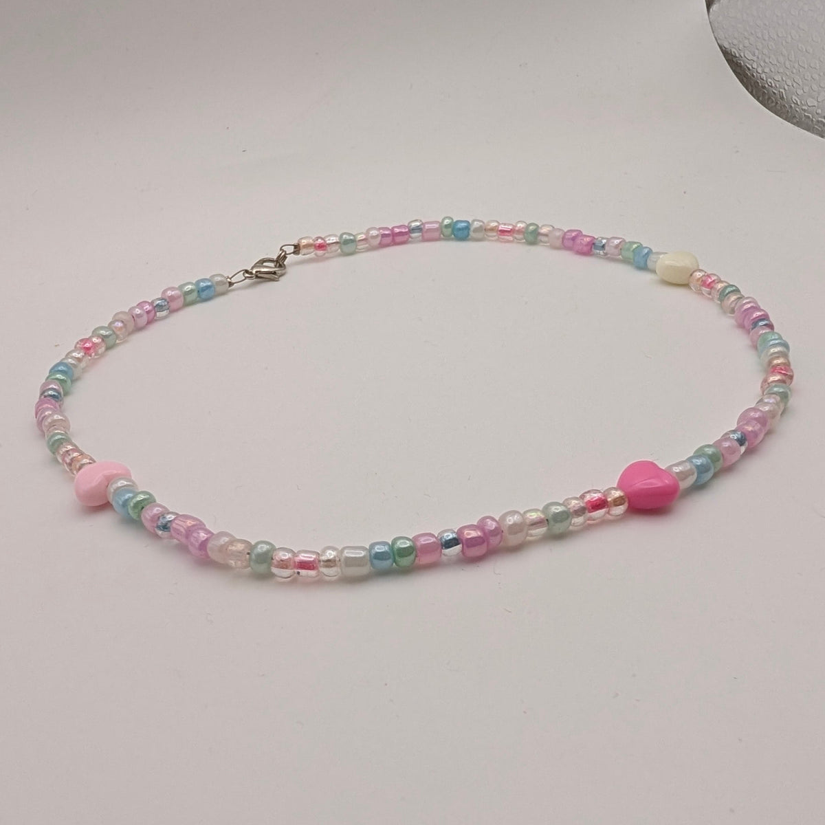 A pastel beaded necklace with pink and blue beads, a small pink heart charm, and a silver clasp, displayed on a light gray surface.