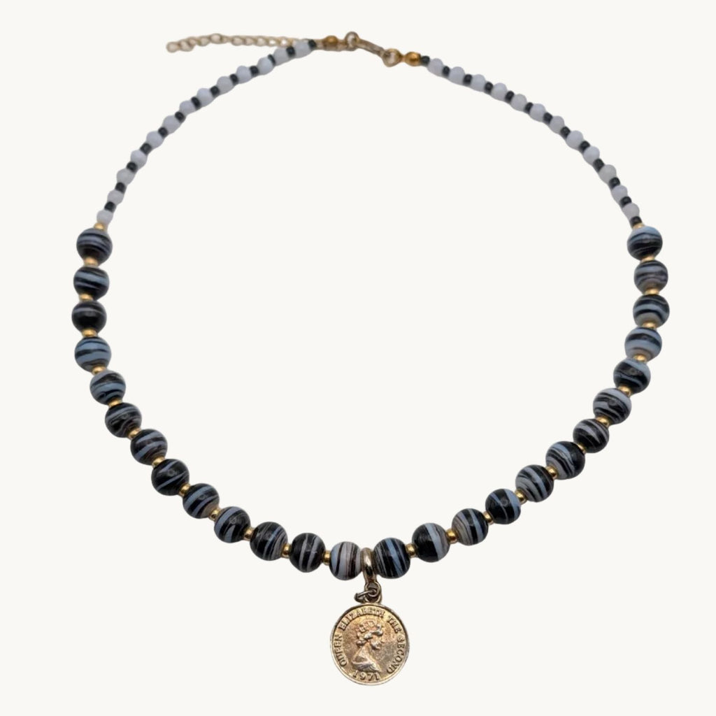 A beaded necklace with dark blue-gray and gold striped beads, featuring a small gold tone coin-style charm in the center, displayed on a white surface.