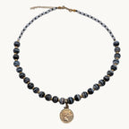 A beaded necklace with dark blue-gray and gold striped beads, featuring a small gold tone coin-style charm in the center, displayed on a white surface.
