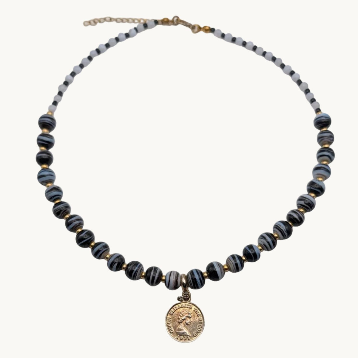 A beaded necklace with dark blue-gray and gold striped beads, featuring a small gold tone coin-style charm in the center, displayed on a white surface.