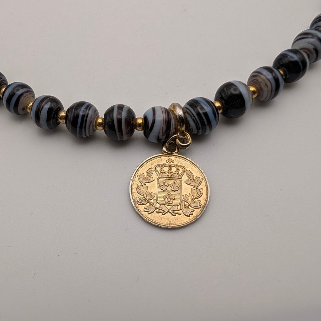 A close-up of a beaded necklace featuring dark, marbled beads with black and gray swirls, accented by small gold spacers and a round gold tone coin-style pendant.