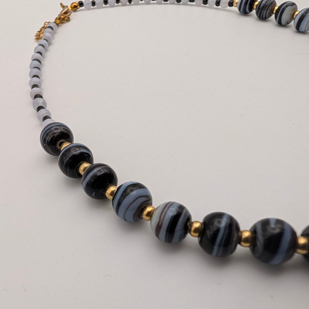 A beaded necklace featuring glossy black and gray striped glass beads, light gray round beads, and small gold tone spacer beads, displayed on a neutral light gray background.