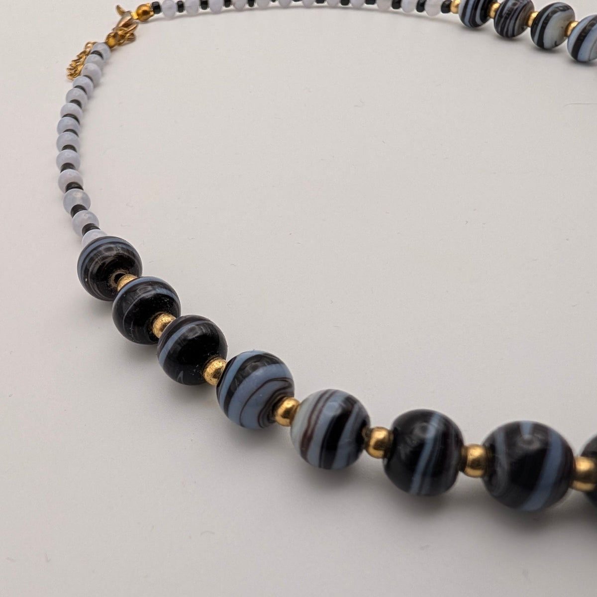 A beaded necklace featuring glossy black and gray striped glass beads, light gray round beads, and small gold tone spacer beads, displayed on a neutral light gray background.