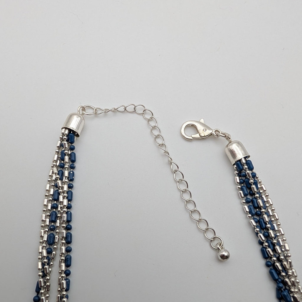 A multi-strand beaded tassel necklace with dark blue and silver beads, featuring a silver lobster clasp and extension chain, displayed on a light gray background.