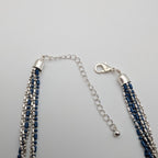 A multi-strand beaded tassel necklace with dark blue and silver beads, featuring a silver lobster clasp and extension chain, displayed on a light gray background.