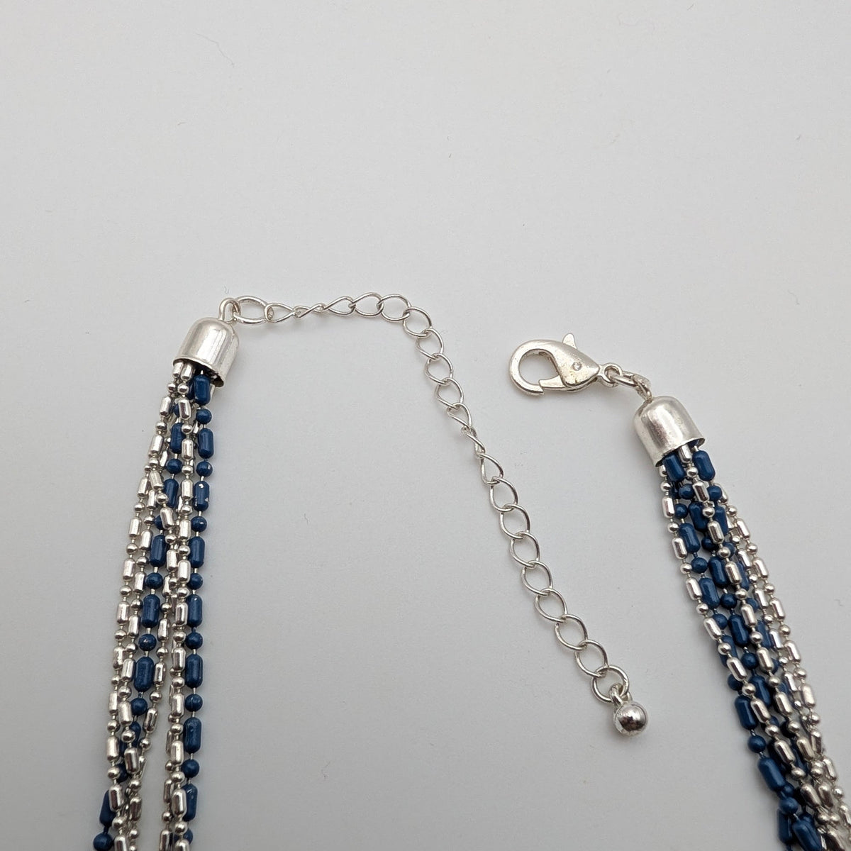 A multi-strand beaded tassel necklace with dark blue and silver beads, featuring a silver lobster clasp and extension chain, displayed on a light gray background.