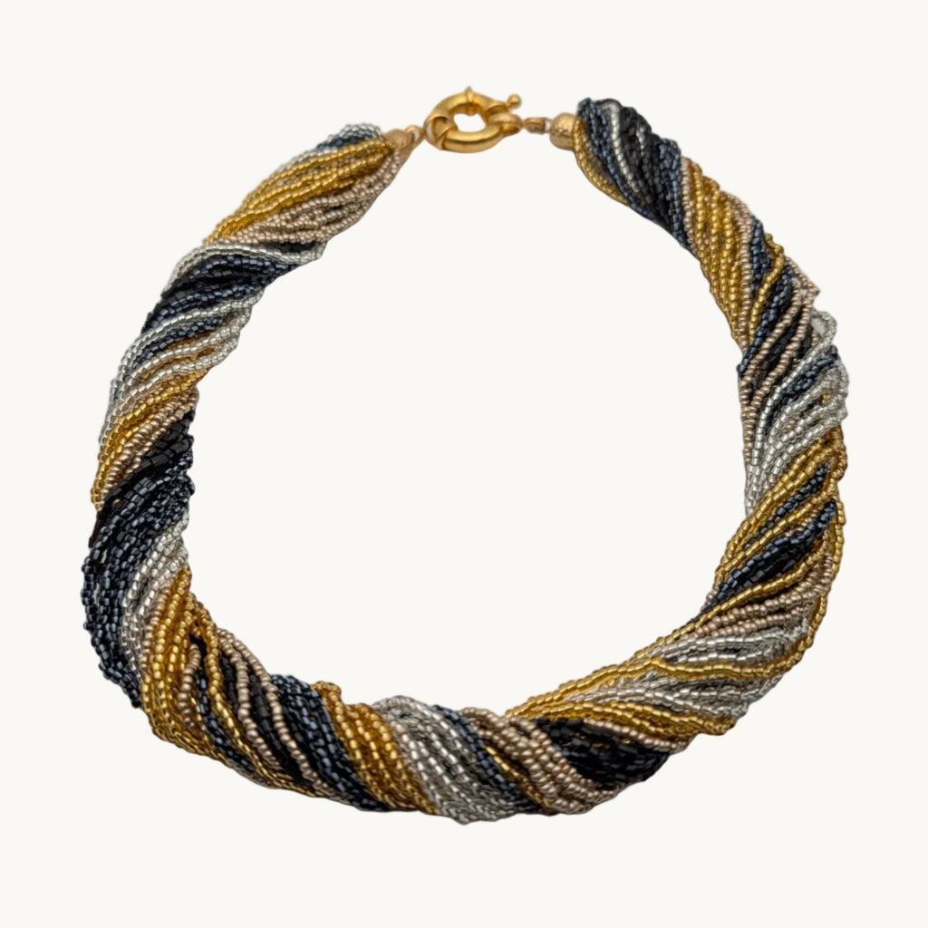 A twisted beaded necklace with a gold clasp, featuring strands of gold, black, and silver beads, displayed on a neutral light gray background.