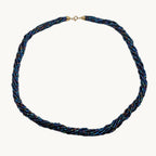 A twisted braided necklace made of blue, black, and gold metallic threads, featuring a small gold clasp at the top, displayed on a plain light gray background.