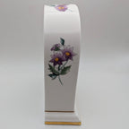 A white ceramic table clock with a curved top and base, decorated with purple flowers and green leaves, standing on a light gray background.