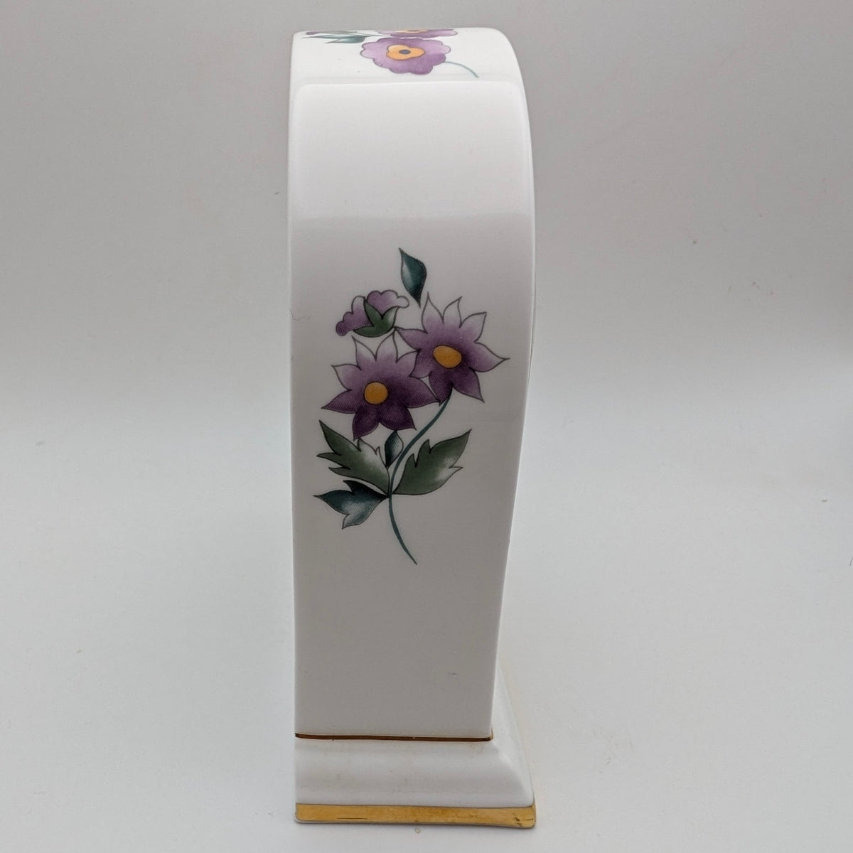 A white ceramic table clock with a curved top and base, decorated with purple flowers and green leaves, standing on a light gray background.