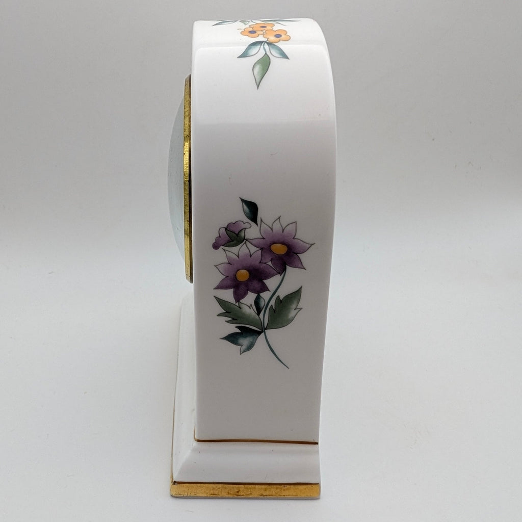 A white porcelain mantel clock with purple flowers, green leaves, and gold trim, shown from a side profile view on a light gray background.
