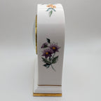 A white porcelain mantel clock with purple flowers, green leaves, and gold trim, shown from a side profile view on a light gray background.