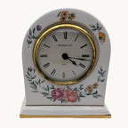 A Wedgwood table clock with a white porcelain body, gold trim, and delicate floral illustrations in pink, yellow, and green, set against a plain white background.