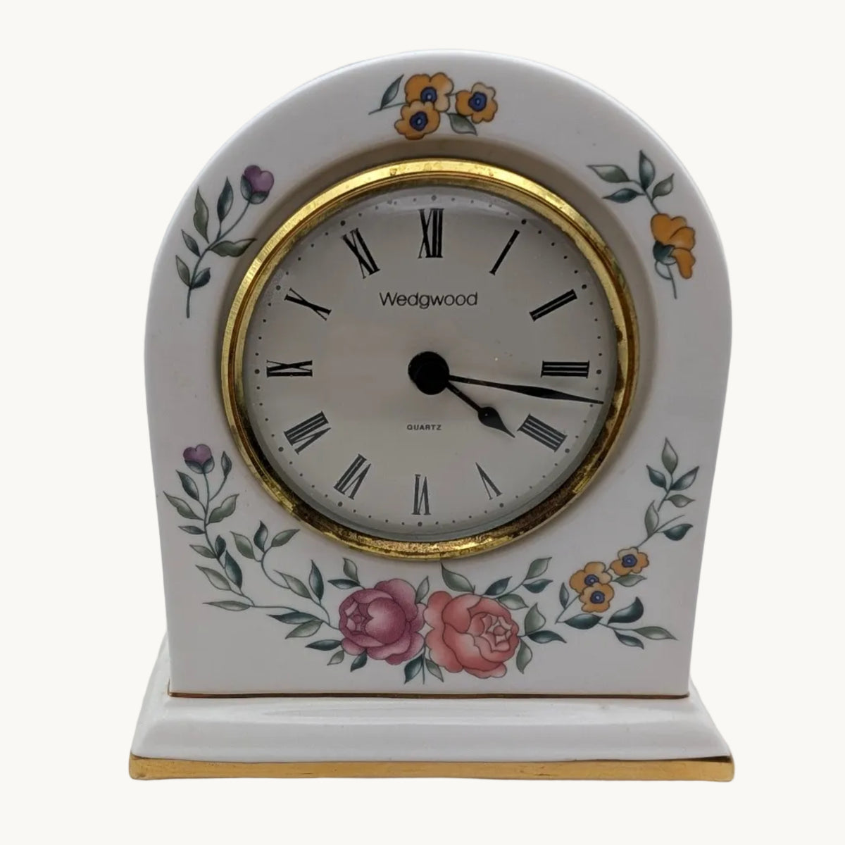 A Wedgwood table clock with a white porcelain body, gold trim, and delicate floral illustrations in pink, yellow, and green, set against a plain white background.