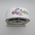 A white ceramic table clock with a floral hand-painted design featuring purple flowers, yellow blossoms, and green leaves, set against a plain light gray background.