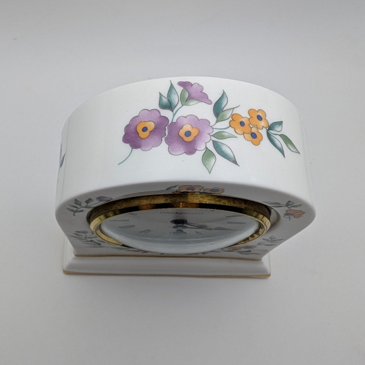 A white ceramic table clock with a floral hand-painted design featuring purple flowers, yellow blossoms, and green leaves, set against a plain light gray background.