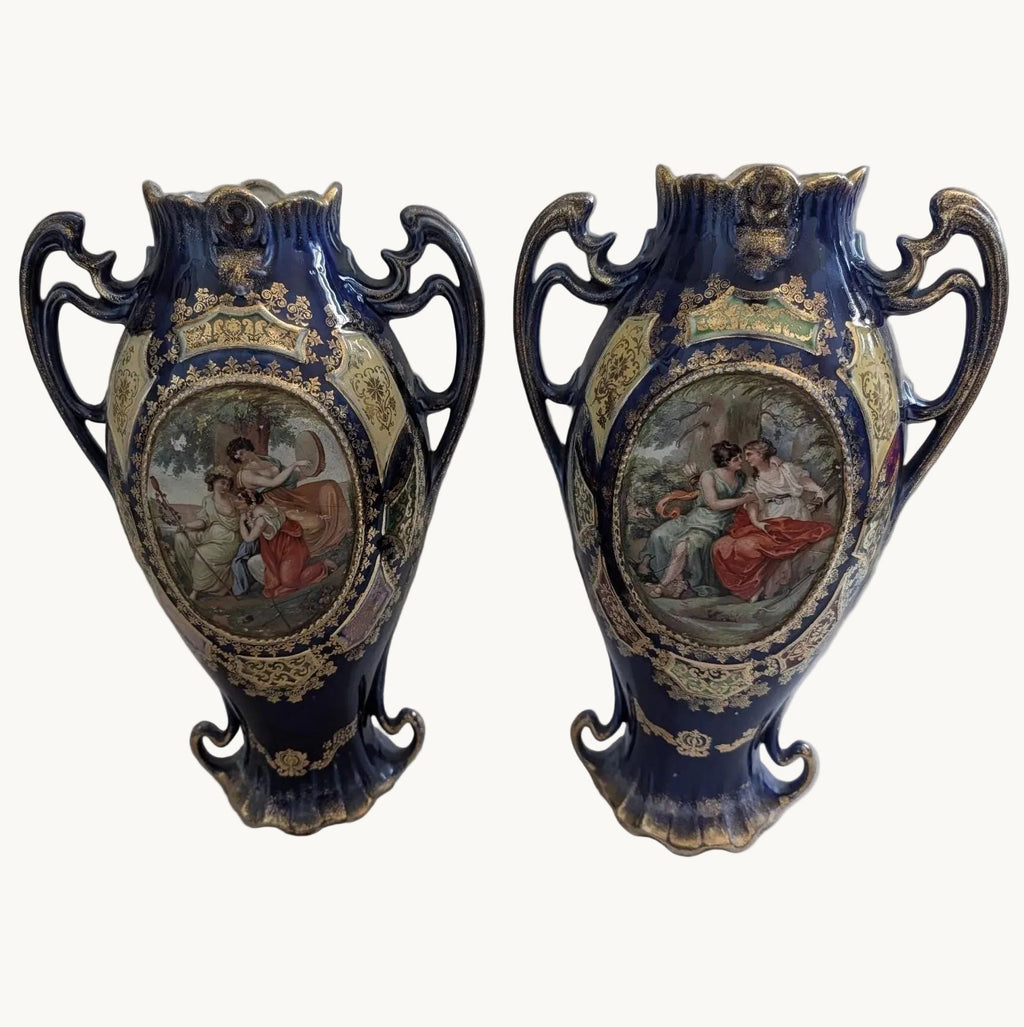 A pair of ornate dark cobalt blue porcelain vases with gold gilding and central painted classical scenes, displayed on a light surface against a grey background.